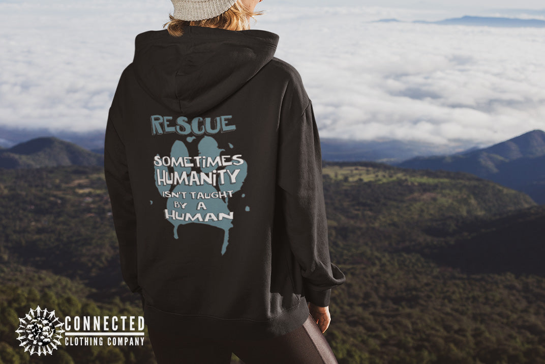 Model wearing Black Show Humanity Unisex Hoodie at the mountains that reads "Rescue. Sometimes humanity isn't taught by a human" - sweetsherriloudesigns - Ethically and Sustainably Made - 10% donated to the Society for the Prevention of Cruelty to Animals